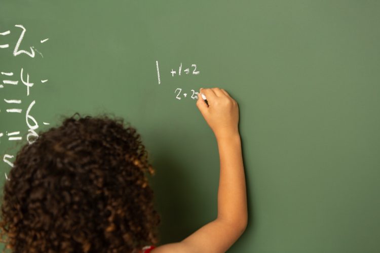 schoolgirl solving mathematics calculations on green board in classroom at school