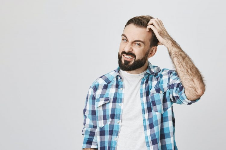 Studio shot of mad bearded male roughly scretching his head and expressing confusion, over gray