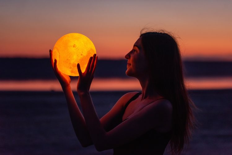 A young woman holds the full moon in her hands against the backdrop of a red sunset. Astrology