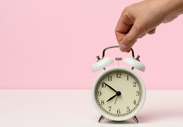 Female hand holds a white metal alarm clock on a white table, time is ten to eight in the morning
