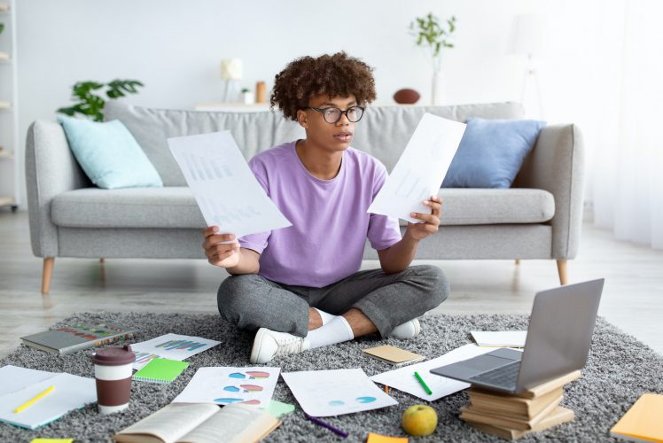 Focused black teen sitting on floor among scattered documents, using laptop to write coursework