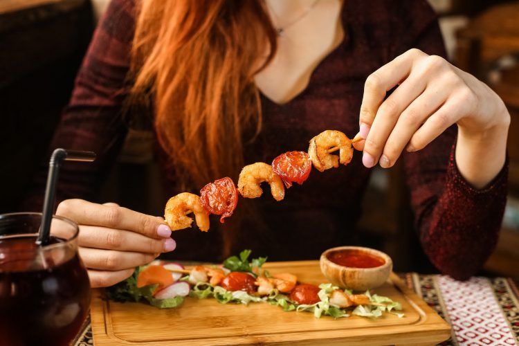 side view woman eating shrimp on a skewer with vegetables and sauce on a stand