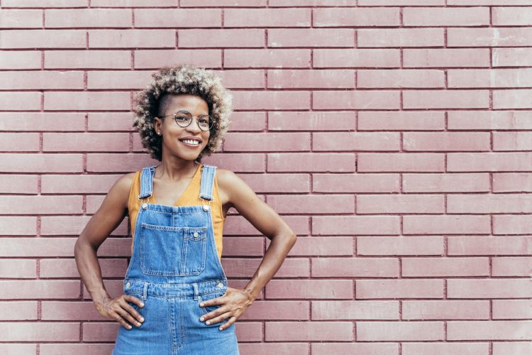 Confident smiling African American Woman with casual bib jeans overall over a brick wall