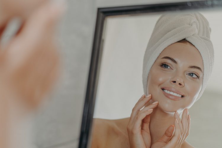 Adorable woman looks at her reflection in mirror, touches healthy glowing smooth skin