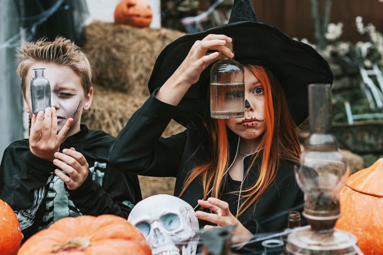 girl in a witch costume and a boy in a skeleton costume looks through the glass lantern on the porch