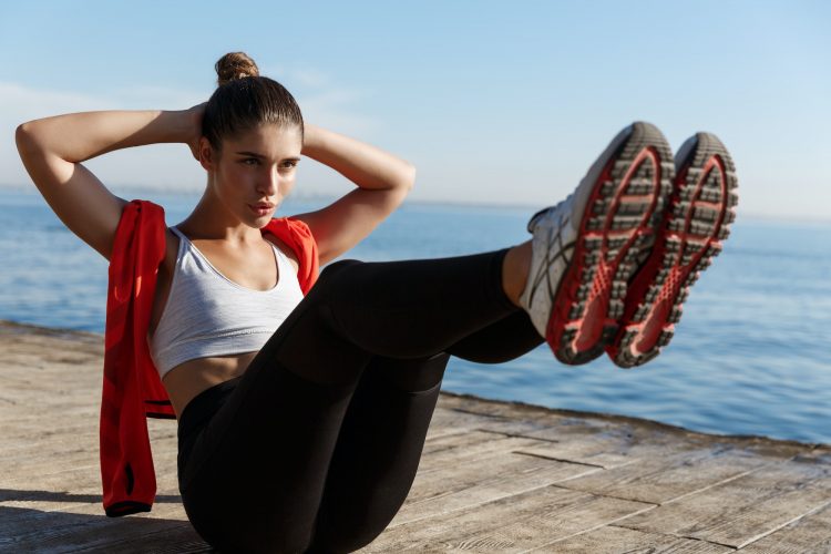 Young confident sportswoman doing crunches on a pier, training abdominal abs, working out on seaside