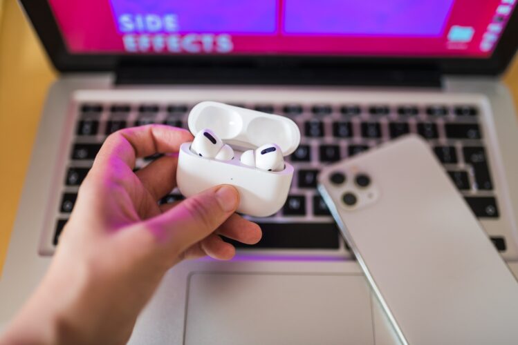Hand holding a wireless headphones open case above a macbook laptop and iphone