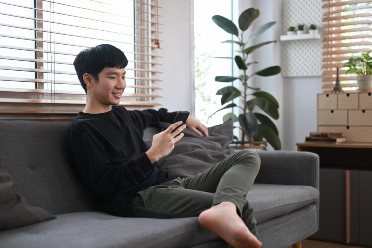 Handsome man resting on couch and typing text message on mobile phone.