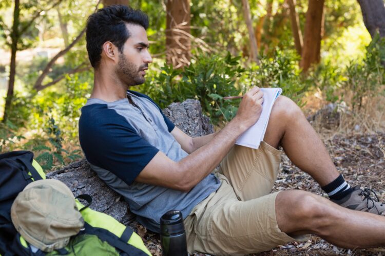 Man writing on notepad while resting on tree trunk