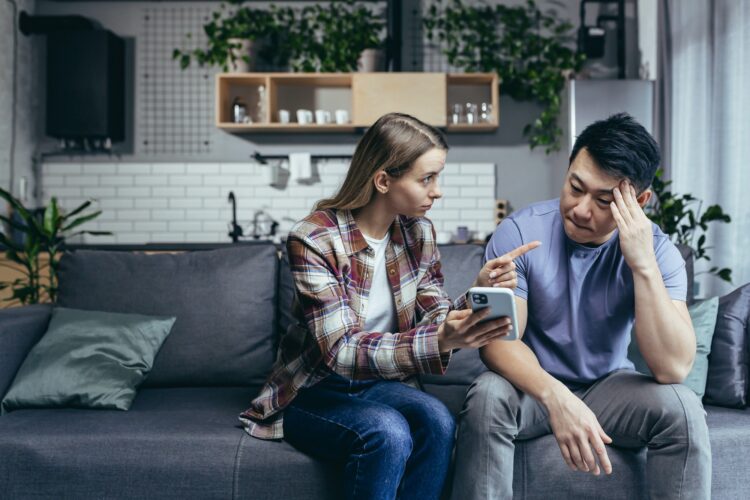 Married couple, man and woman, sitting on the couch at home and arguing over the phone, jealousy