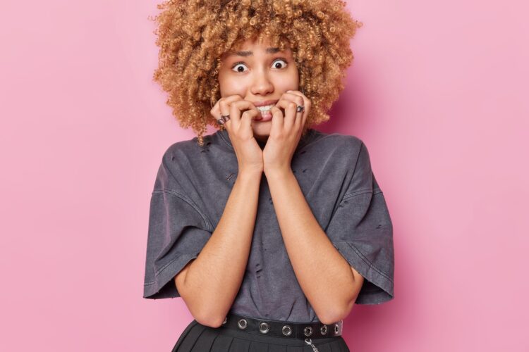 Waist up shot of nervous curly haired woman bite finger nails feels nervous looks anxious at camera