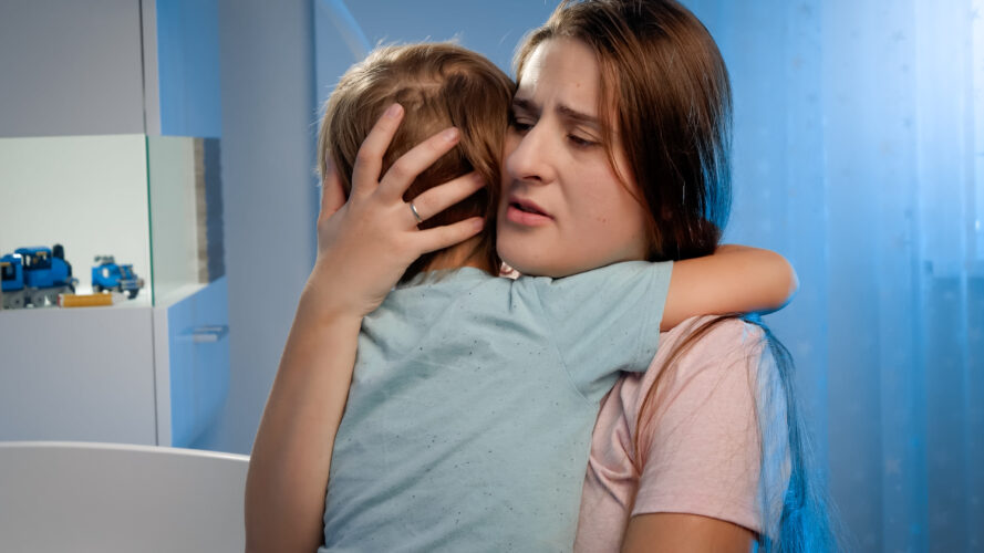 Portrait of young loving mother soothing and caressing scared little boy in children room at night