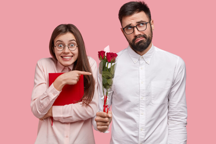 Positive young European woman carries red textbook, points at awkward bearded man in white shirt who