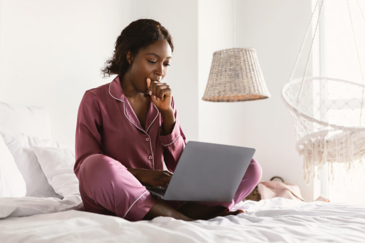 Sleepy African Freelancer Lady Using Laptop And Yawning In Bedroom