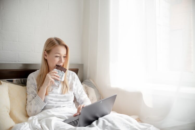 Young happy woman freelance working in bed and eating chocolate. Work from home and self employment