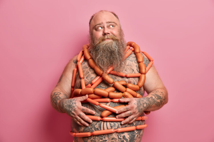 Food addiction and bad nutrition concept. Plump bearded man stands wrapped in sausages has big fat a