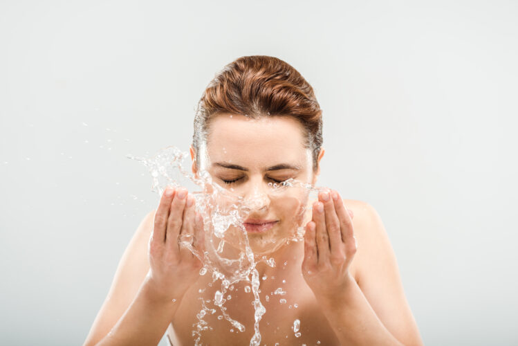 naked woman with closed eyes washing face on grey