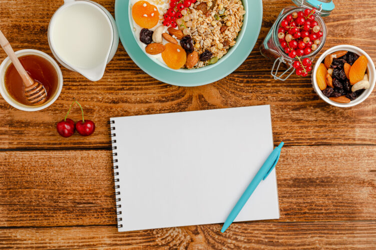 White blank of food diary, healthy breakfast of granola with fruits, milk and honey on wooden table.