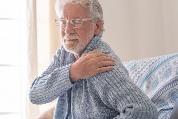 70s senior man sitting at home on sofa suffering for muscle strain or arthritis on shoulder