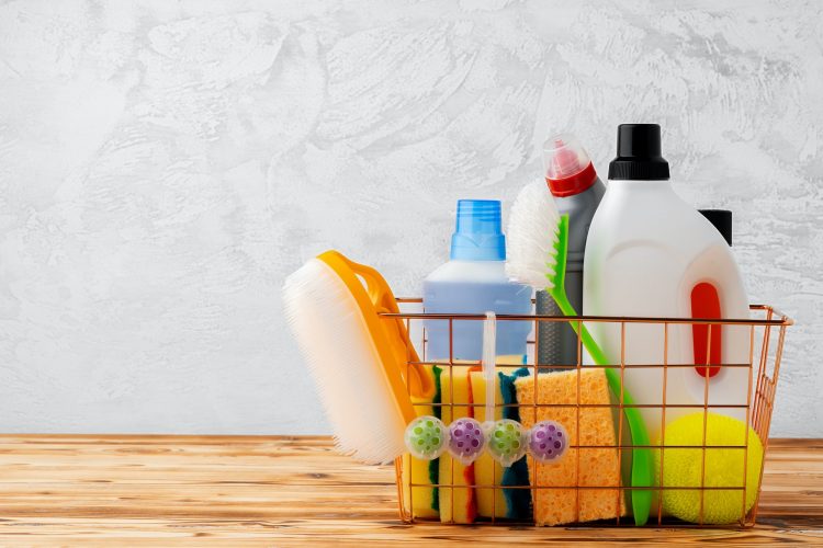 Cleaning tools and chemicals in basket on wooden table against gray background