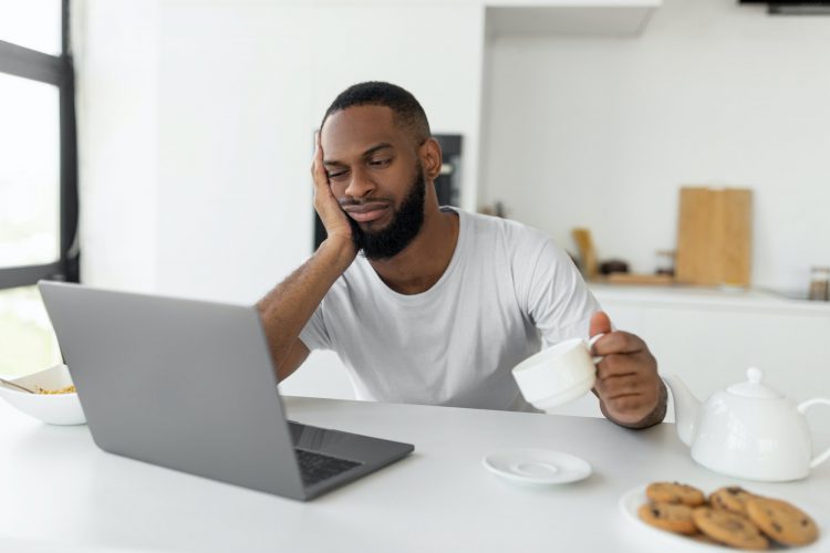 Tired black man using laptop sitting at kitchen table