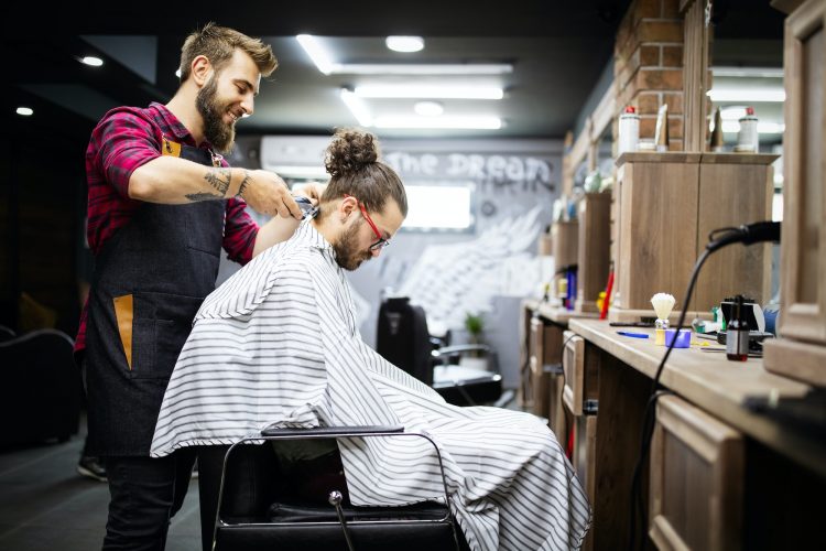 Young bearded man getting haircut by hairdresser while sitting in chair at barbershop
